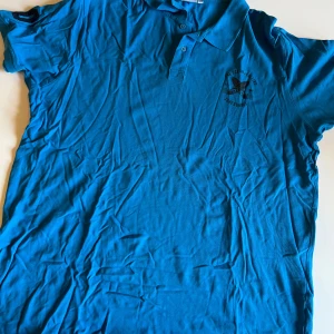 Blå pikétröja från Hangten XXXL - Snygg blå pikétröja från Hangten i storlek XXXL. Tröjan har klassisk krage, knäppning med två knappar och tryck med surfbrädor och texten 'The Ocean is Calling Surf Boards' på bröstet. Perfekt för en chill och sportig look.