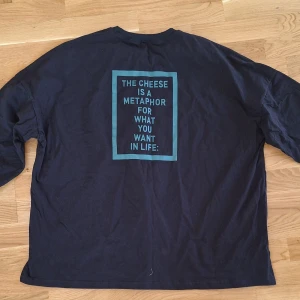 Svart långärmad tröja med tryck Oversized XL - Svart långärmad tröja i bomull med blått tryck på ryggen: 'The cheese is a metaphor for what you want in life' och texten 'cheeses cracker' längs ena sidan. Tröjan har rund hals och muddar vid ärmslut. Perfekt för dig som gillar unika statement-plagg.