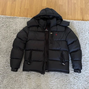 Svart pufferjacka från Polo Ralph Lauren - Svart pufferjacka från Polo Ralph Lauren med huva, dragkedja och två fickor framtill. Jackan har en röd broderad logga på bröstet och är vadderad för extra värme. Perfekt för kalla dagar och har en klassisk, sportig look. Jackan är helt ny använd en gång. Säljer för att den var för stor