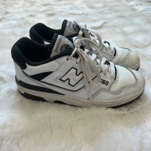 New Balance 550 vita sneakers - Säljer ett par klassiska New Balance 550 sneakers i vitt med svarta detaljer. Skorna har snörning, rund tå och platt sula. Ovandelen är i skinn och textil, med tydlig New Balance-logga på sidan och 550-tryck. Perfekta för dig som gillar retrostil och streetwear.