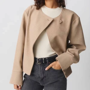 Beige kort kappa från Gina Tricot - Kort beige kappa med minimalistisk design och rund halsringning. Jackan har en enkel knäppning med en synlig knapp upptill och vida ärmar. Tillverkad i ullblandning för en mjuk och stilren look. Perfekt för lager-på-lager och snygg till jeans.