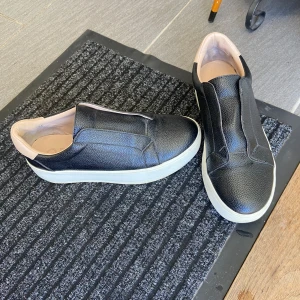 Svarta slip-on sneakers med vit sula,40 - Snygga svarta slip-on sneakers från Officine Creative med vit platt sula och rund tå. Skorna är gjorda i strukturerat skinn och har beige detaljer vid hälen och insidan. Perfekta för en clean och stilren look.storlek 40. Hämtas i norsborg botkyrka.