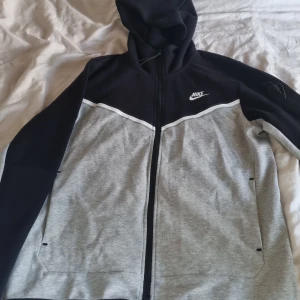 Nike tech hoodie och byxor - Nike hoodie i svart och ljusgrå med vit logga på bröstet. Tröjan har huva,  och två fickor framtill. Materialet är mjuk bomull och polyester, säljer dessa kläder här på grund av storleken helt ny oanvänt och redo för en ny ägare. Vid frågor om kläderna skriv gärna.