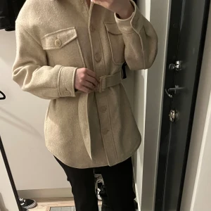 Kappa Nakd - Snygg beige overshirt med bälte i midjan och stora bröstfickor med knappar. Jackan har lång ärm, klassisk krage och knäppning framtill. Materialet är mjukt och känns som ull, perfekt för lager-på-lager. Stilren och enkel att matcha med olika outfits.