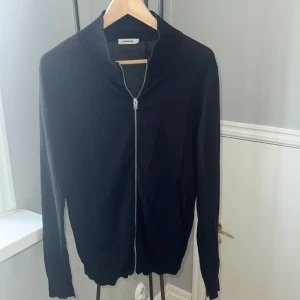 Svart kofta med dragkedja J.Lindeberg - Svart kofta från J.Lindeberg i storlek S. Tröjan har en stilren design med ribbad krage och hel dragkedja framtill. Materialet är mjukt och stickat, perfekt för lager-på-lager. Långärmad och med fickor på sidorna för extra chill vibe.