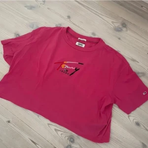 Rosa croppad t-shirt Tommy Jeans - Snygg rosa croppad t-shirt från Tommy Jeans med tryckt logga i flera färger på bröstet. T-shirten har korta ärmar och en relaxed fit, perfekt för en chill och trendig look.