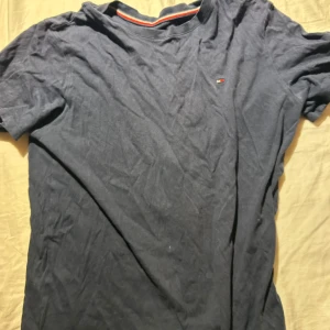Mörkblå t-shirt från Tommy Hilfiger - Mörkblå t-shirt från Tommy Hilfiger med liten broderad logga på bröstet. Klassisk rund halsringning med röd och vit randig detalj på insidan av kragen. Tillverkad i mjuk bomull och har korta ärmar. Perfekt basic-plagg med snygg passform. Storlek 160-175cm (14-16 år)