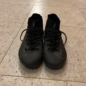 Svarta fotbollsskor med snörning - Säljer ett par svarta fotbollsskor med högt skaft och snörning. Skorna har mönstrad ovandel och dobbar under sulan för bra grepp på planen. Snygg, slimmad design som passar perfekt för dig som vill sticka ut på matchen.