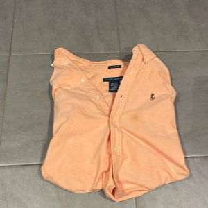 Persikofärgad skjorta från Ralph Lauren - Snygg och trendig skjorta i bra skick! Märkt med strl 4, vilket motsvarar ca XS/S. Perfekt för dig som vill ha en stilren skjorta som passar både till vardag och mer uppklädda tillfällen.  Jag är 182 cm och den sitter lite för tight på mig, men passar säkert för någon som är lite mindre. Har två små fläckar (se bild 3 och 4) tvättade skjortan men fläckarna försvann inte. Men annars är den i bra skick. 