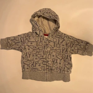 Grå hoodie med gitarrmönster H&M - Grå hoodie från H&M i storlek 62, täckt av svarta gitarrer och små stjärnor. Tröjan har dragkedja framtill, huva och ribbade muddar. Perfekt för små rockstjärnor som vill sticka ut lite extra.