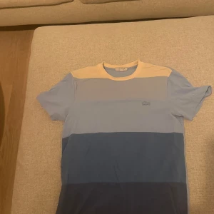 Randig t-shirt från Lacoste i blå toner - Snygg t-shirt från Lacoste med breda horisontella ränder i ljusgul, ljusblå och mörkblå. Klassisk passform med rund hals och korta ärmar. Ikoniska Lacoste-loggan broderad på bröstet. Perfekt för dig som gillar stilrena och fräscha färger.