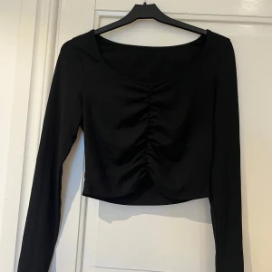 Svart ribbad croppad topp - Säljer en svart långärmad topp med ribbad struktur och snygg rynkdetalj framtill. Modellen är croppad och har en lätt v-ringad halsringning. Perfekt att styla med höga jeans eller kjol. 