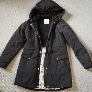 Elvine - Svart parkasjacka från Elvine, M (dam) - Svart parkasjacka från Elvine med huva och stora fickor framtill. Jackan har dragkedja och knappar, samt ett unikt innerfoder med ugglemönster. Perfekt för kyliga dagar med sin längre modell och snygga detaljer.