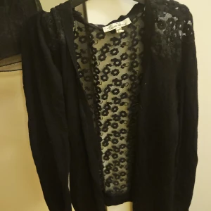 Svart kofta med spets från Miss Snob - Svart kofta från Miss Snob med transparent spets och blommigt mönster framtill och över axlarna. Långärmad och öppen framtill, perfekt för att ge outfiten en extra touch. Materialet är mjukt och spetsen ger en cool och feminin vibe.