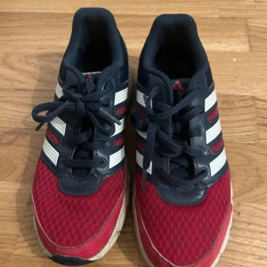 Adidas sneakers i rött och marinblått - Snygga Adidas sneakers med klassiska vita ränder, ovandel i röd mesh och marinblå detaljer. Skorna har snörning och platt sula, perfekt för en sportig look. Materialet är en mix av syntet och textil för bra komfort.