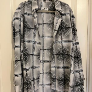 Gråvit rutig skjorta från Ledi - Säljer en gråvit skjorta från Ledi med ett unikt rutigt mönster i svart och blått. Skjortan har lång ärm, klassisk krage och bröstficka. Materialet känns mjukt och har en lätt glans, perfekt för dig som gillar att sticka ut med coola detaljer. Storlek 42 Män. Använd en gång. 