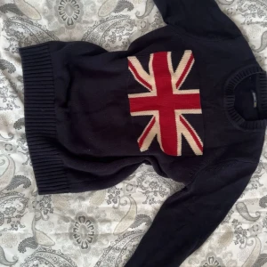 Mörkblå stickad tröja från carved - Snygg mörkblå stickad tröja från Gant med Union Jack-flagga i rött, vitt och blått på bröstet. Tröjan har rund halsringning, ribbade muddar och långa ärmar. Perfekt för dig som söker en clean och old money look 