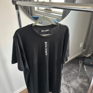 Svart Palm Angels t-shirt med tryck - Svart t-shirt från Palm Angels med stort vitt logotyp-tryck längs ryggen och mindre tryck framtill. Storlek Medium. Nyskick.
