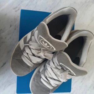 Adidas Campus grå sneakers stl 42 - Snygga Adidas Campus sneakers i grå mocka med klassiska vita ränder och vit sula. Skorna har rund tå, platt sula och snörning. Ikonisk Adidas-logga på plösen och Campus-tryck på sidan. Perfekta för en stilren och avslappnad look.