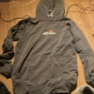 Grå hoodie från Ellesse med logga - Säljer en grå hoodie från Ellesse i storlek S. Tröjan har klassisk huva med vita snören, stor magficka och Ellesse-logga broderad på bröstet. Perfekt för chill dagar eller när du vill ha en enkel och snygg look.