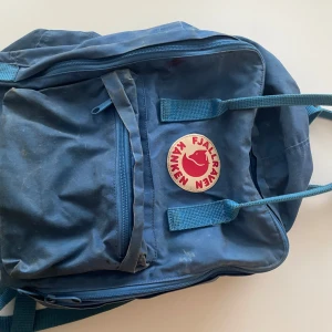 Blå Fjällräven Kånken ryggsäck - Klassisk blå Fjällräven Kånken ryggsäck med ikonisk rund röd logga framtill. Väskan har ett stort huvudfack, en mindre ficka med dragkedja på framsidan och justerbara axelremmar. Materialet är slitstark polyester och nylon, perfekt för skolan eller utflykter.