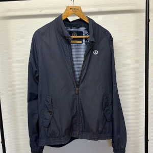 Marinblå vindjacka från Henri Lloyd - Snygg marinblå vindjacka från Henri Lloyd med dragkedja och två fickor framtill. Jackan har ett klassiskt rutigt innerfoder i blått och vitt samt broderad logga på bröstet. Perfekt för vår och höst när du vill ha en stilren look.