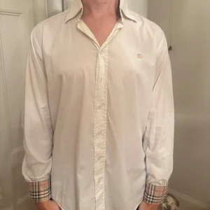 Vit Burberry skjorta med rutiga detaljer - Snygg vit skjorta från Burberry med klassisk krage och knappar framtill. Ärmarna är långärmade och har den ikoniska Burberry-rutiga detaljen vid mudden. Perfekt för dig som gillar stilrena plagg med exklusiv touch. Köpt på second hand butiken Napoli Centrale