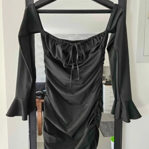 Svart offshoulder klänning med volang - Svart kortklänning med offshoulder-modell och långa ärmar med volang vid ärmslut. Klänningen har snörning vid bysten och rynkade detaljer längs sidorna för en snygg passform. Perfekt för dig som gillar trendiga och stilrena plagg.