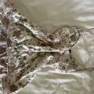 Blommig kortklänning från Passport - Supersöt kortklänning från Passport i 100% bomull med blommigt mönster i vitt/beige, grönt och lila/rosa. Klänningen har smala axelband, knyt i ryggen och volangkant nertill. 