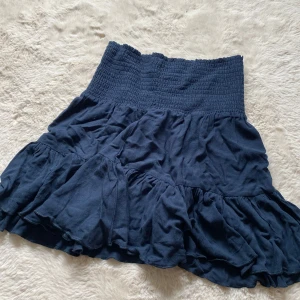 Mörkblå volangkjol med smock - Supersöt mörkblå kortkjol med bred smock i midjan och flera volanglager. Kjolen har en luftig och flowy känsla, perfekt för varma dagar. Enkel att matcha med både toppar och sneakers för en chill look.