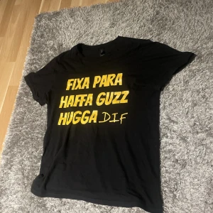 Svart t-shirt med gul text - Svart t-shirt med tryck i gult: 'FIXA PARA HAFFA GUZZ HUGGA DIF'. Klassisk passform och rund hals. Perfekt statement-plagg för dig som vill sticka ut. Materialet känns mjukt och skönt mot huden.