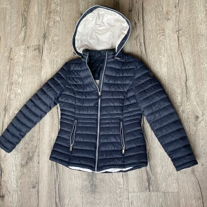 Mörkblå pufferjacka från Saki - Mörkblå höst/vårjacka från Saki med snygg figursydd design Luva finns men går att ta av samt på igen med dragkedja. Jackan har två fickor med dragkedja i fram och en innerficka. Super fin kvalitet och en snygg jacka i allmänhet men storleken är inte rätt för mig därför är den nu till salu. Nypriset låg runt 2000kr därav mitt pris på 1000kr men kan absolut diskutera priset om nödvändigt.