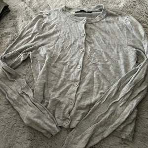 Grå kofta från 157 med knappar - En enkel och stilren grå kofta från 157 i storlek M. Koftan har rund halsringning, långa ärmar och knäppning framtill med matchande knappar. Tillverkad i mjukt bomullsmaterial, perfekt för lager-på-lager under kyligare dagar.