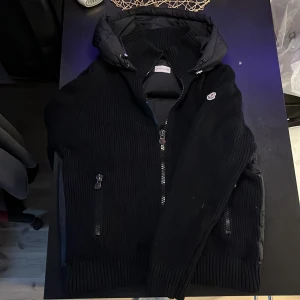 Svart dunväst med huva från Moncler - Svart dunväst från Moncler med stickade sidor och ribbade detaljer. Storlek L men passar M. Västen har en quiltad baksida, dragkedja framtill, två fickor med dragkedja och en stor huva. Perfekt för lager-på-lager för en stilren look. Perfekt inför det kalla vädret och endå se fly ut. Den fångar blickar från tjejer😉