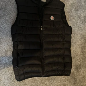 Svart dunväst från Moncler - Svart dunväst från Moncler med quiltad design och dragkedja framtill. Västen har en klassisk Moncler-logga på bröstet och en stilren, sportig look. Perfekt för lager-på-lager och passar till många outfits.