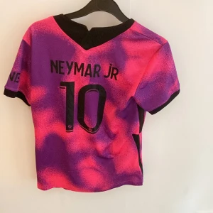 PSG Neymar Jr fotbollströja rosa/lila - Säljer en PSG fotbollströja med Neymar Jr och nummer 10 på ryggen. Tröjan har ett unikt rosa och lila mönster, svarta detaljer och klubbmärke framtill. Tillverkad i lätt och ventilerande material, perfekt för träning eller match. Priset kan förhandlas vill bara få den såld