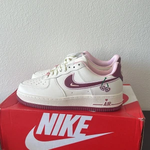 Nike Air Force 1 Cherry Love sneakers - Unika Nike Air Force 1 sneakers i vitt och vinrött med rosa detaljer. Skorna har broderade körsbär och hjärtan, samt hjärtformad Nike-logga på hälen. Platt sula, snörning och lågt skaft. Tillverkade i skinn med rosa innerfoder och vinröd yttersula. Perfekta för dig som vill sticka ut.