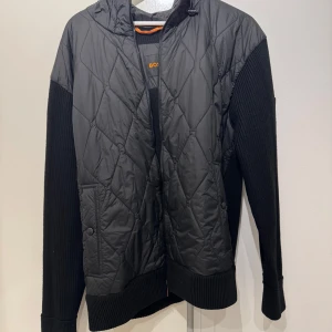 Boss orange cardigan jacka (nyskick) - Säljer en svart jacka från BOSS orange med quiltad framsida och huva. Ärmar och rygg är stickade för en schysst kontrast. Jackan har dragkedja och två fickor framtill. Perfekt för dig som vill ha en stilren och modern look. Jackan är i fortfarande i nytt skick, ca använd 2 gånger vilket syns på lappen vid insidan 