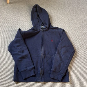 Ralph Lauren Polo (junior) - Marinblå hoodie - Marinblå hoodie från Polo Ralph Lauren med dragkedja framtill och klassisk huva. Tröjan har lång ärm och en liten röd broderad logga på bröstet. Tillverkad i mjuk bomull, perfekt för chill dagar eller när du vill ha en enkel och stilren look.