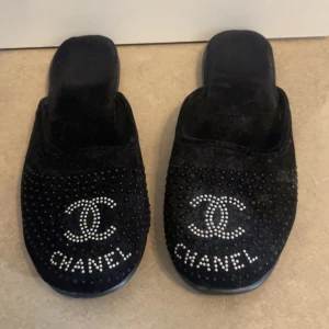 Svarta Chanel slip-ins med strass - Svarta slip-in tofflor från Chanel med mjuk sammet och dekorerade med strassdetaljer samt den ikoniska Chanel-loggan framtill. Skorna har platt sula och rund tå, perfekta för en lyxig och chill look.