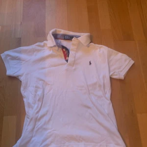 Vit pikétröja från Polo Ralph Lauren - Vit pikétröja från Polo Ralph Lauren med klassisk krage och korta ärmar. Snygga blåvita ränder på insidan av kragen och detaljer i rött och blått vid knäppningen. Broderad logga på bröstet. Tillverkad i mjuk bomull för en skön känsla.