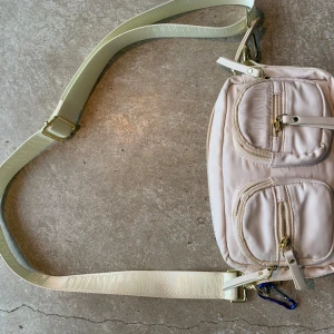 Beige axelväska från Poppy Lissiman - Snygg beige axelväska från Poppy Lissiman med bred justerbar rem, två framfickor med dragkedja och guldfärgade detaljer. Ny utan prislapp, men kvitto och jag medföljer. Nypris 1400:-