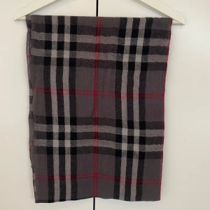 Rutig grå halsduk med röda detaljer - Stilren halsduk i grått med klassiskt rutigt mönster i svart, vitt och röda linjer. Tunn och mjuk känsla, perfekt att slänga över axlarna. Avslutas med små fransar i kanterna. Passar dig som gillar en tidlös vibe.