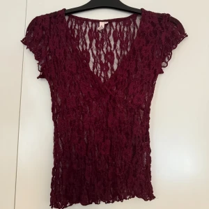 Vinröd spetsblus från Nelly XS - Säljer en vinröd blus från Nelly i storlek XS. Blusen är helt i spets med v-ringning och korta ärmar. Den har en genombruten, romantisk look och är figurnära i modellen. Perfekt för dig som gillar detaljer och vill sticka ut med en snygg topp.