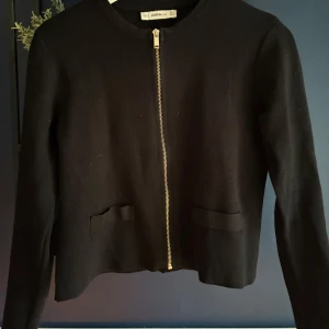 Svart kofta med dragkedja från Zara - Stilren svart kofta från Zara med rund halsringning och snygg guldfärgad dragkedja framtill. Koftan har två dekorativa fickor och är tillverkad i ett stickat material som känns mjukt och bekvämt. Perfekt för lager-på-lager och enkel att matcha.