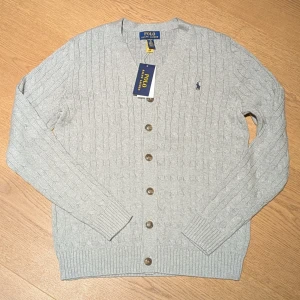 Ny grå kabelstickad kofta Polo Ralph Lauren, junior kille  - Ny/oanvänd kabelstickad grå kofta från Polo Ralph Lauren med klassisk v-ringning och knappar framtill. Liten broderad logga på bröstet och ribbade muddar vid ärmslut och nederkant. Perfekt för lager-på-lager och snygg till jeans. Storleken är junior kille XL (motsvarande 170/176). Nypris är 1450 kr, jag säljer den för 1000 kr.