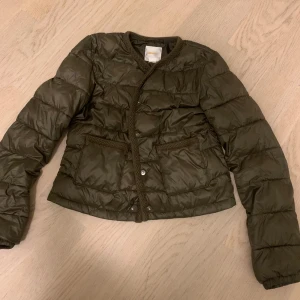 Höstjackan  - Snygg olivgrön pufferjacka med flätade detaljer längs kanterna och vid fickorna. Jackan har tryckknappar framtill och två stora fickor. Perfekt för dig som vill ha en stilren och trendig look under kyliga dagar.