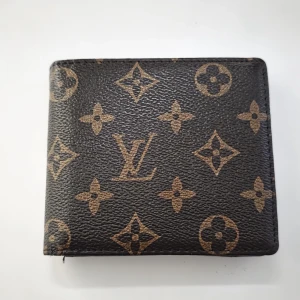 Plånbok i monogram - Snygg plånbok med klassiskt monogrammönster i brunt och beige. Plånboken är i skinn och har flera kortfack samt myntficka. Perfekt för dig som vill ha en ikonisk accessoar med lyxig känsla.