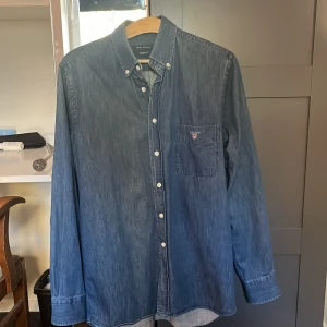 Jeansskjorta GANT - Exklusiv blå jeansskjorta från GANT i indigo twill. Klassisk button-down krage, vita knappar och bröstficka med broderad GANT-logga. Skjortan är tillverkad i Portugal och har en relaxed passform. Perfekt för en chill och stilren look. Ny pris omkring 2000kr.