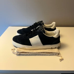 Valentino Garavani sneakers i mocka - Snygga sneakers från Valentino Garavani i mörkblå mocka med vita skinnpaneler och coola nitar på sulan. Klassisk rund tå, svarta skosnören och vit platt sula. Märkeslogga på plösen och insidan. Perfekta för dig som gillar lyxiga detaljer.